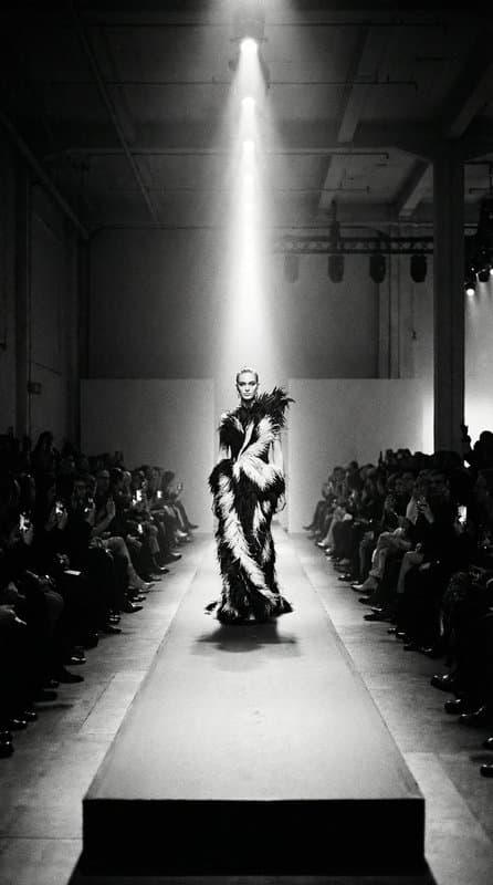 A single model in a feathered look walking a runway between seated rows