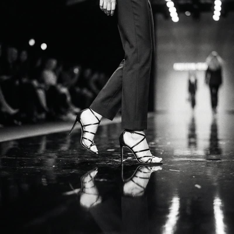 Ankle and heel mid-stride on a wet mirrored runway, a figure receding behind